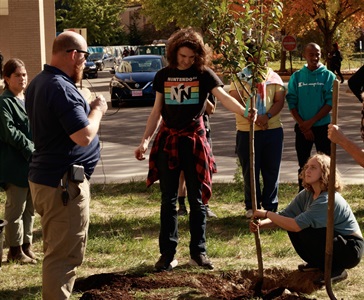 Two young people with the Sustainability Club at CSCC steady a planted tree while other people watch.