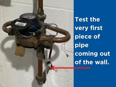 Photo of an indoor water meter and pipes. A red arrow points to the first piece of water service line coming out of the wall, showing where to test the pipe material