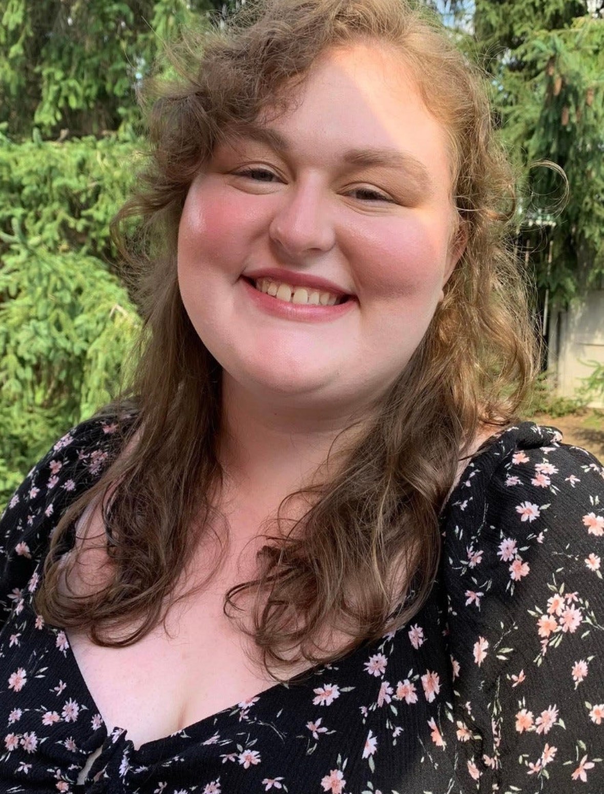 A young woman with wavy, light brown hair is smiling brightly while standing outdoors in front of green trees. She is wearing a black dress with a pink and white floral pattern and has a natural, warm expression. Teh sunlight highlights part of her face and hair. 
