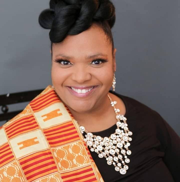 A smiling woman wearing a black dress adorned with a statement pearl necklace and matching earrings. She has an elegant updo hairstyle and is draped in a vibrant Kente cloth featuring traditional gold, orange, and red patterns. The background is a plain dark gray, keeping the focus on her radiant expression and cultural attire. 