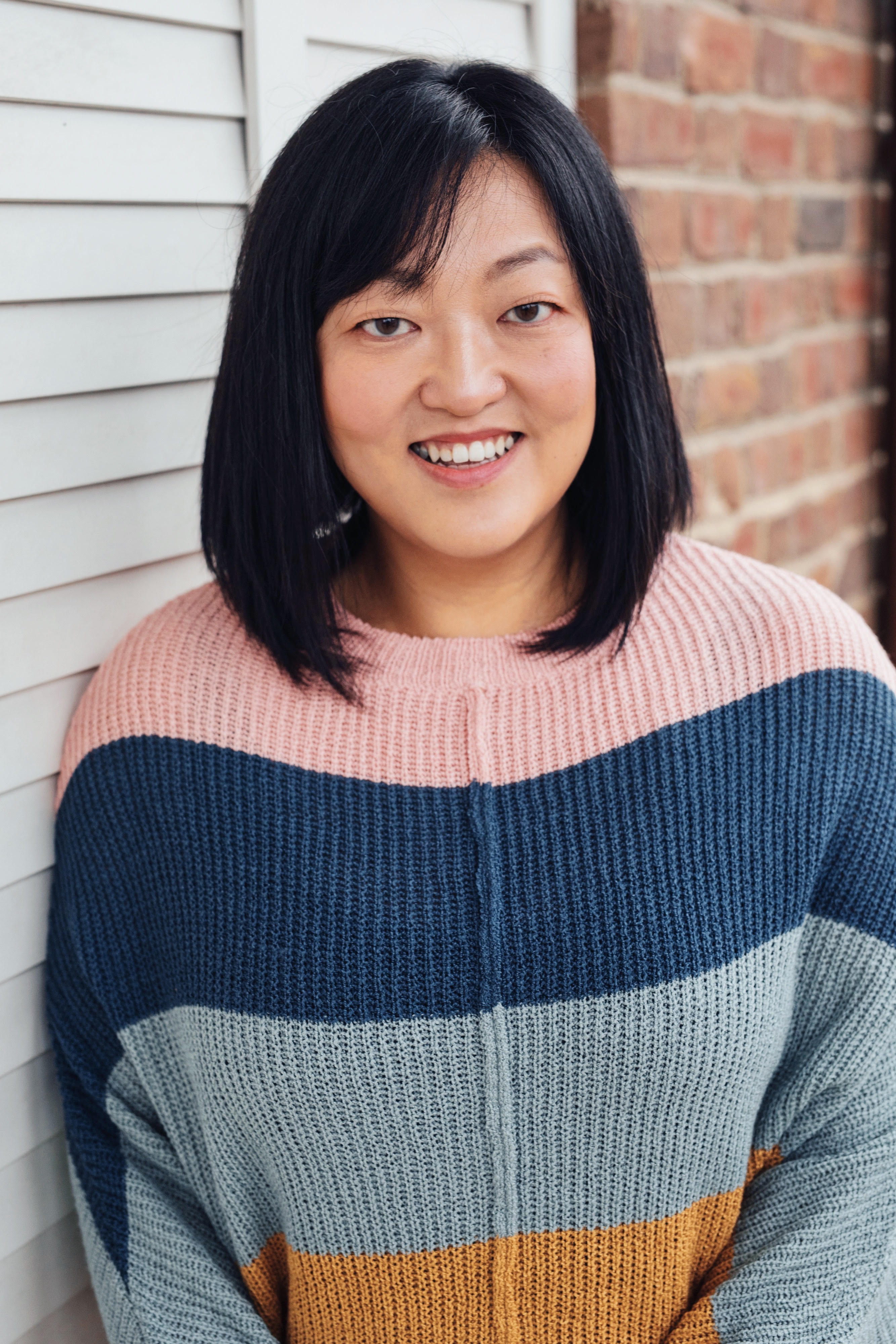 A smiling woman with straight black hair wearing a color-blocked knit sweater in pink, navy blue, light gray, and mustard yellow. She is standing in front of a white paneled wall and a brick wall. 