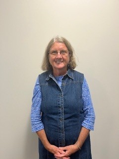 Woman with shoulder-length light brown hair wearing glasses, a blue patterned shirt, and a denim vest, standing and smiling in front of a plain light-colored wall. 