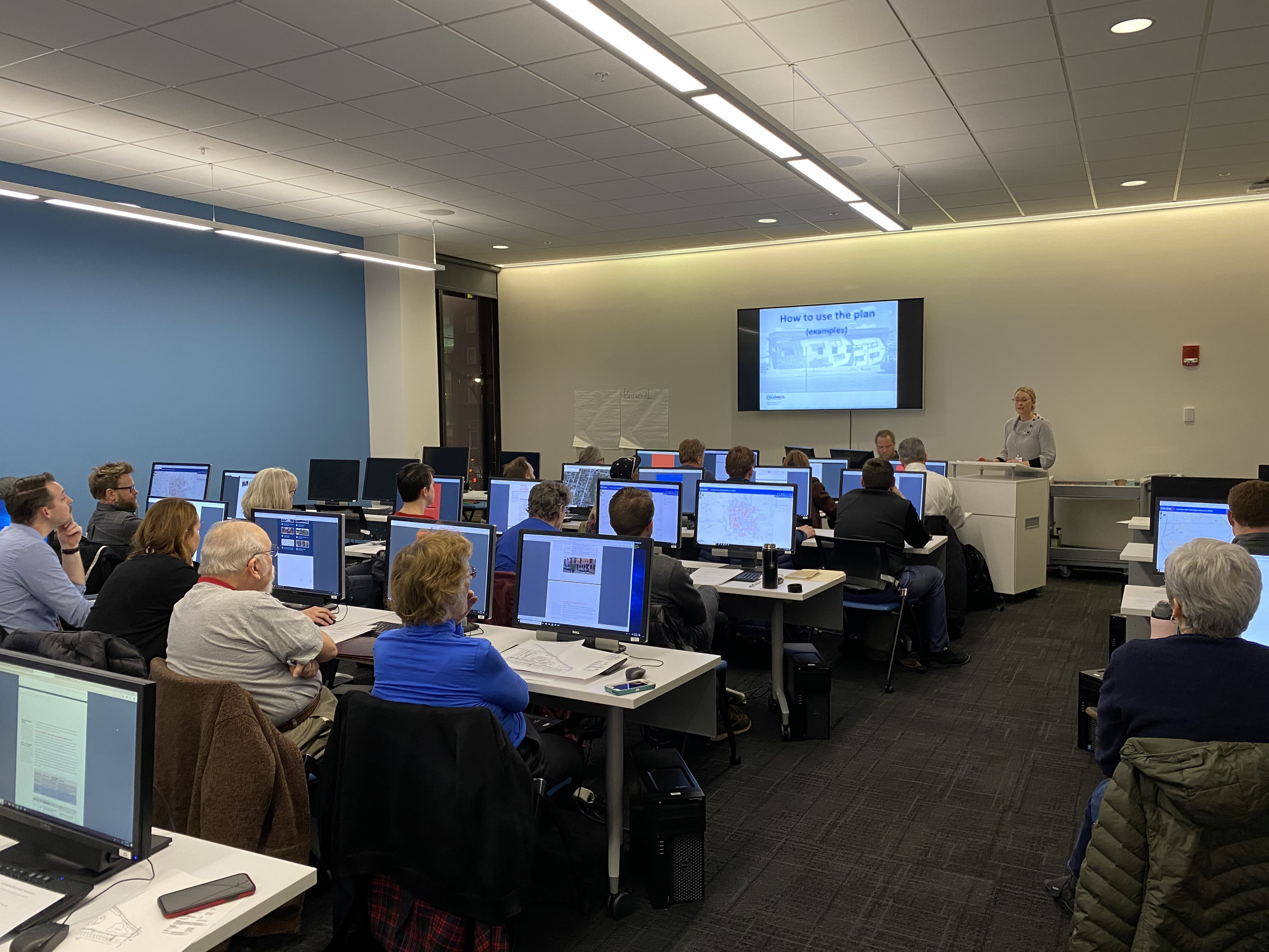 A person stands at a lectern in the front of a computer classroom with people sitting at every computer, facing the front. On the wall a screen has the words, How to use the plan (examples)