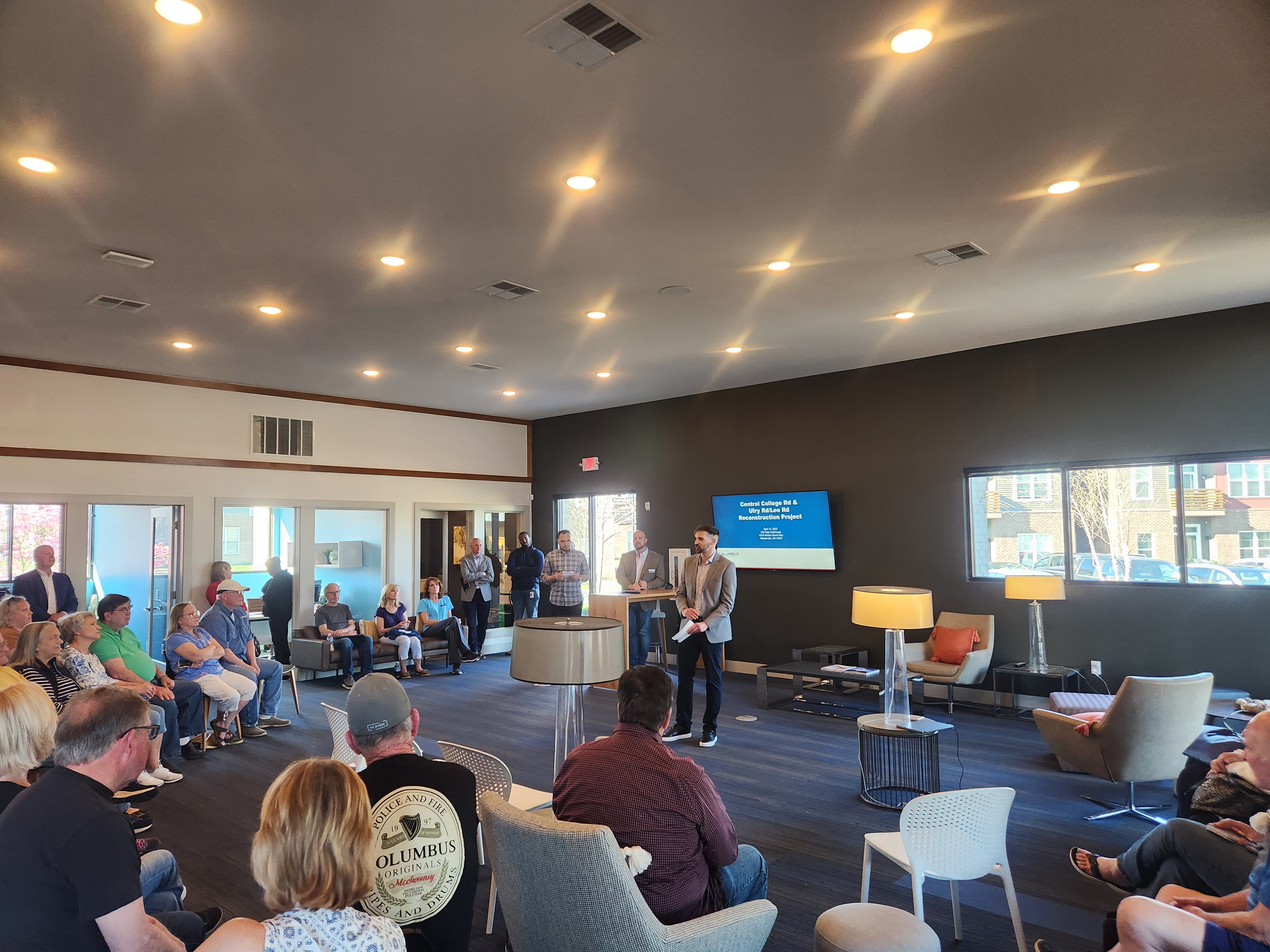 A man in a gray suit coat and black pants stands in the front of a room full of people sitting in different chairs. A screen behind him says, Central College Rd. & Ulry Rd./Lee Rd. Reconstruction Project